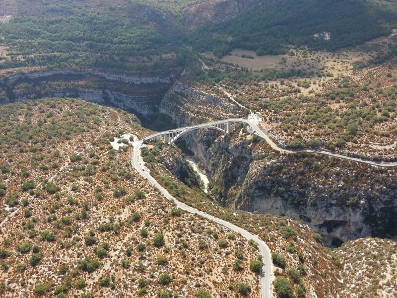 Vue aérienne d'un pont en arc traversant un profond canyon rocheux dans un paysage aride et montagneux.