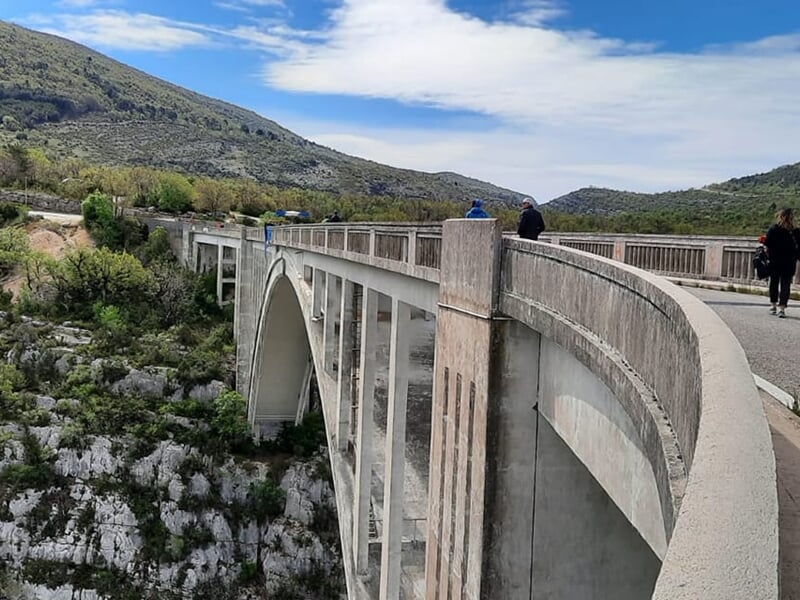 Pont en arc vu de côté avec des personnes marchant dessus, entouré de collines et de végétation verte.