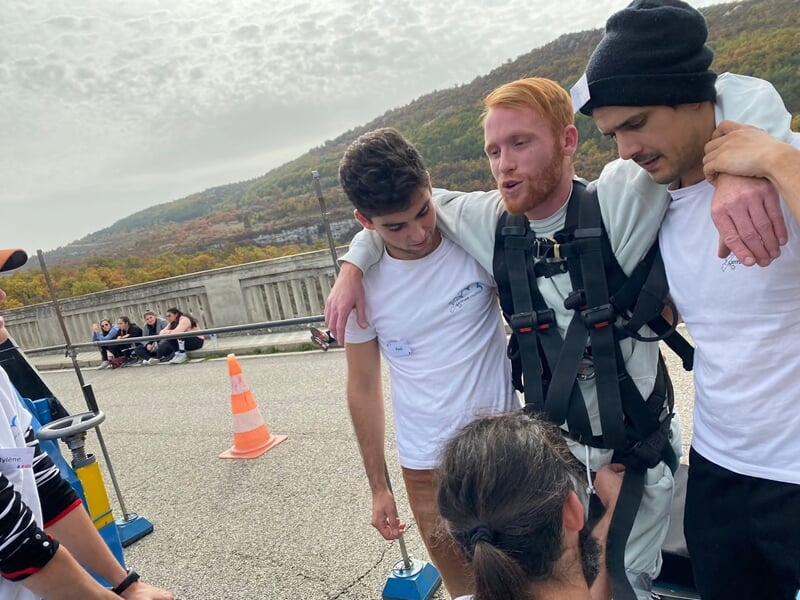 Groupe de jeunes aidant un homme équipé pour un saut à l'élastique sur un pont, ambiance conviviale.