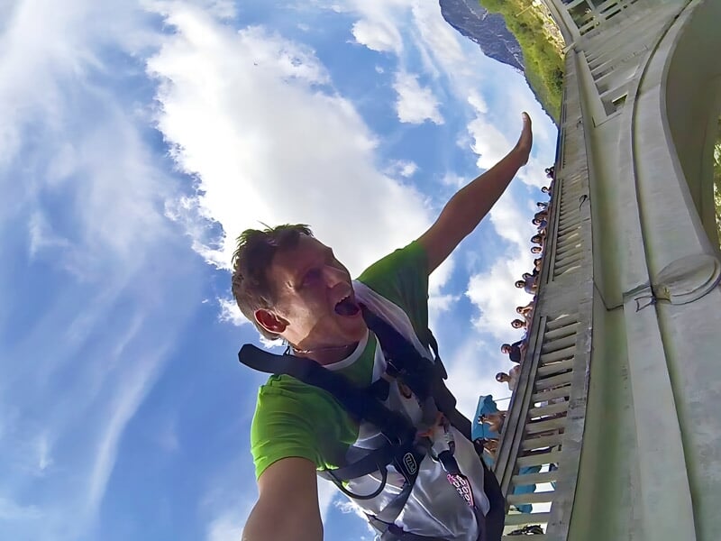 Homme en train de sauter à l'élastique depuis un pont, bras écartés, ciel bleu et spectateurs en arrière-plan.