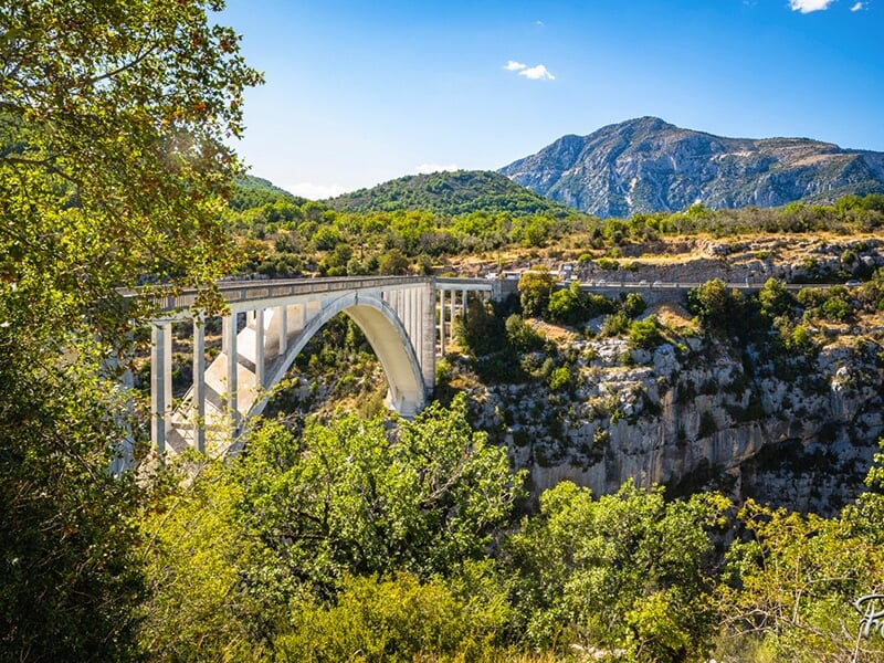 Un pont en arc en béton blanc traversant un canyon verdoyant avec une montagne en arrière-plan sous un ciel bleu dégagé.