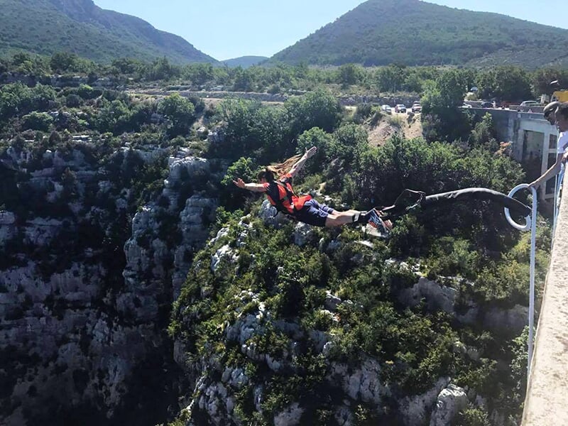 Une femme avec un gilet rouge effectuant un saut à l'élastique depuis un pont, les bras écartés, au-dessus d’un canyon entouré de végétation et de montagnes.