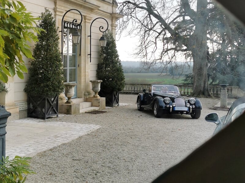 Voiture ancienne noire garée devant un bâtiment classique avec des arbres et une terrasse en pierre.