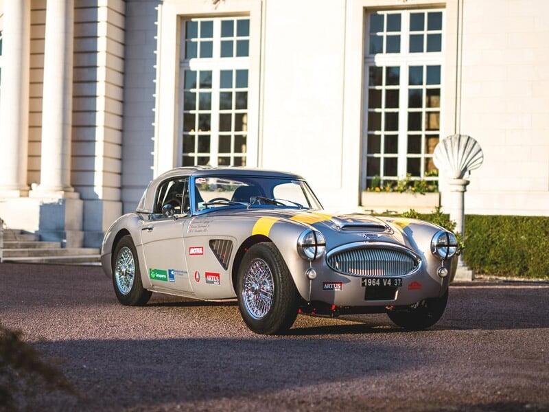 Voiture de course vintage grise avec des autocollants, garée devant un bâtiment ancien ensoleillé.
