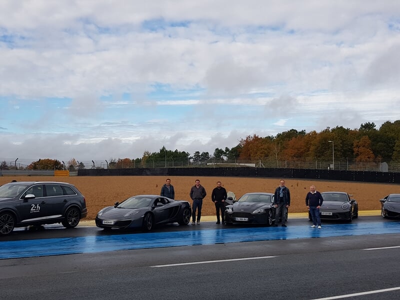 Groupe de personnes debout devant plusieurs voitures de sport garées sur une piste de course.