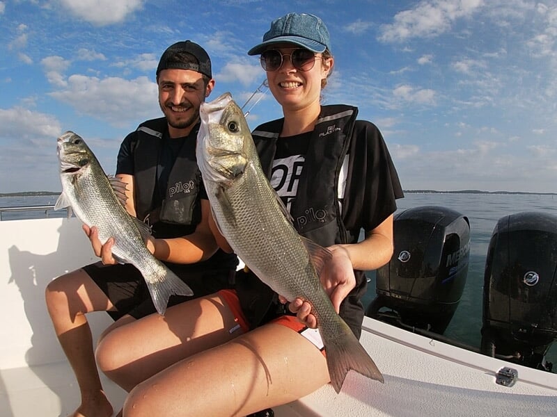 Deux pêcheurs souriants sur un bateau tenant chacun un gros poisson fraîchement pêché en mer sous un ciel clair.