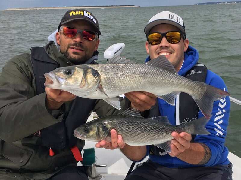 Deux hommes en tenue de pêche sur un bateau exhibent deux gros poissons fraîchement capturés lors d'une sortie en mer.