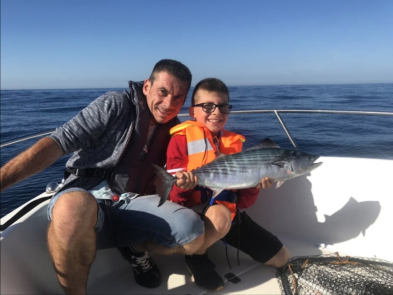 Homme et garçon souriant, tous deux tenant un poisson, le garçon portant un gilet de sauvetage orange, ciel dégagé et mer calme en arrière-plan, filet de pêche à côté d'eux.