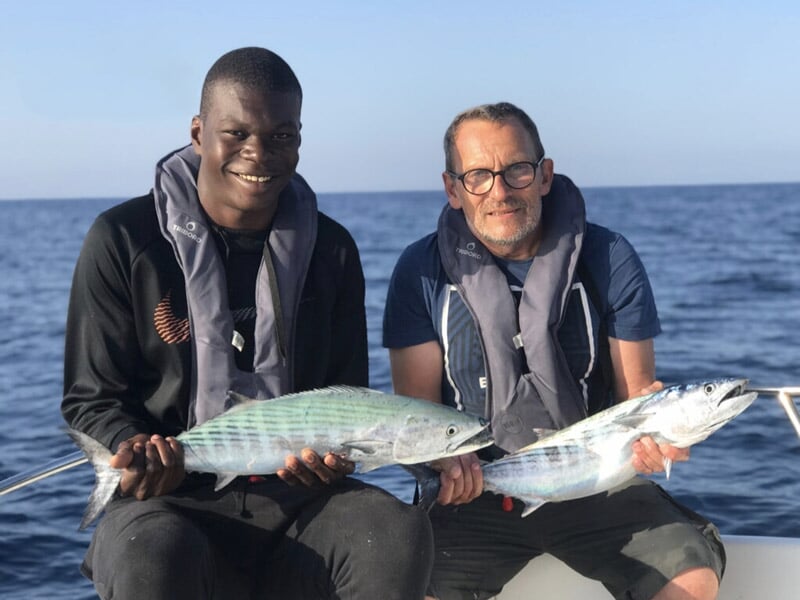 Deux hommes souriants avec des poissons rayés, un jeune homme en noir et un homme plus âgé avec des lunettes, mer bleue et ciel clair.