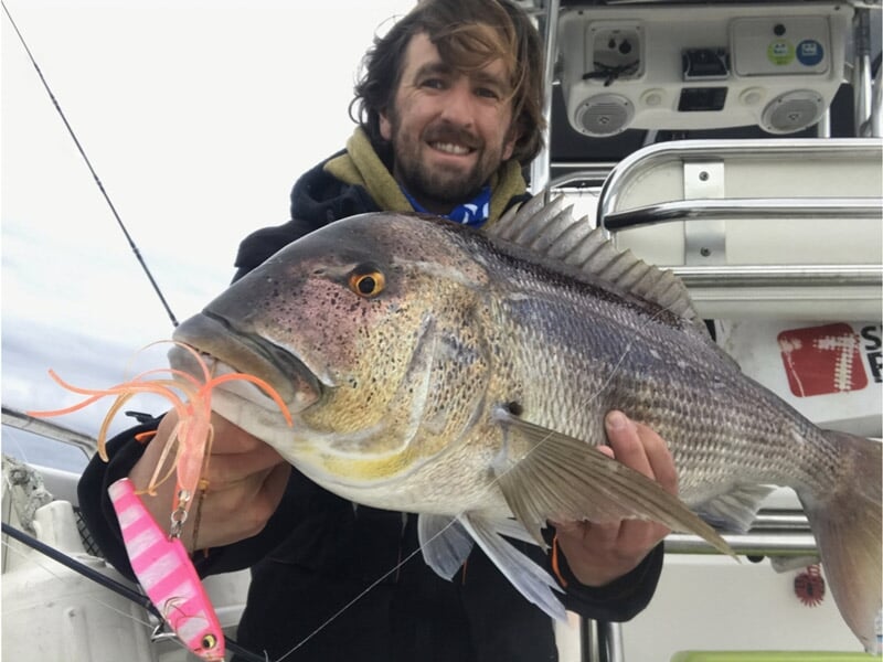 Homme souriant tenant un gros poisson sur un bateau de pêche, appât rose et orange dans la bouche du poisson, bateau équipé de matériel de pêche en arrière-plan. Journée nuageuse en mer. Pêcheur en ma