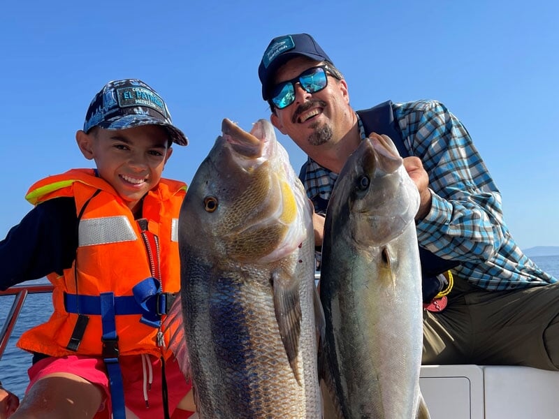 Garçon souriant avec gilet de sauvetage orange et homme en chemise à carreaux, tenant de grands poissons argentés et marron, en mer par ciel dégagé