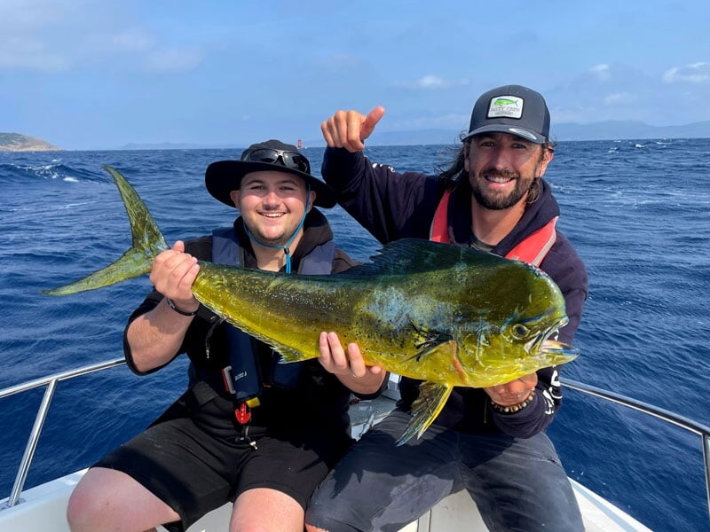 Deux hommes en vêtements de pêche, l'un avec un chapeau noir, souriant et tenant un poisson doré à longue nageoire, sur une mer agitée