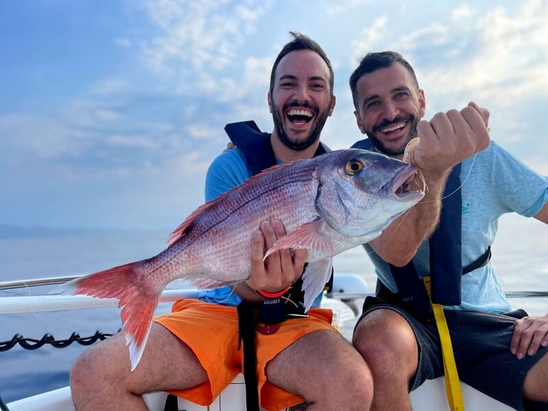 Deux hommes en gilets de sauvetage, l'un riant aux éclats, tenant un poisson rose et blanc avec des reflets argentés, évoquant joie et complicité
