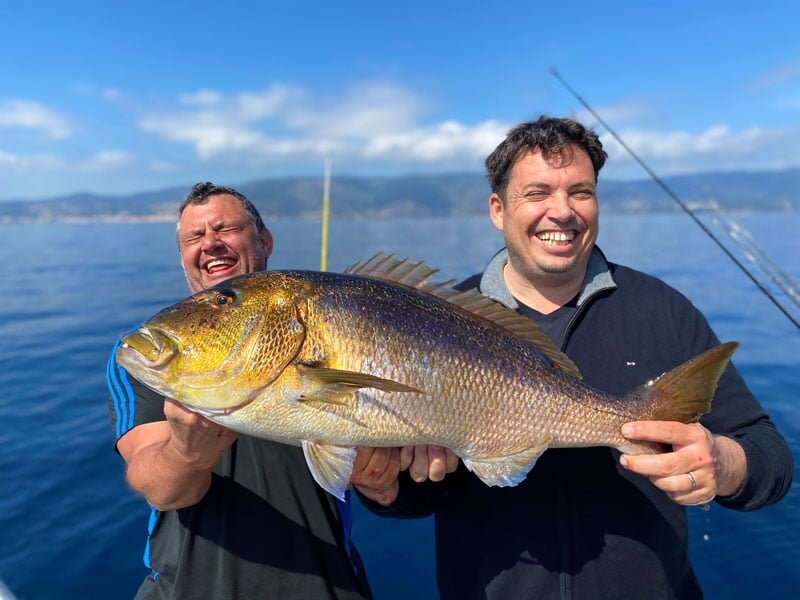 Deux hommes en tenue sportive, riant et tenant un gros poisson doré à grosses écailles, avec la mer et le littoral en arrière-plan