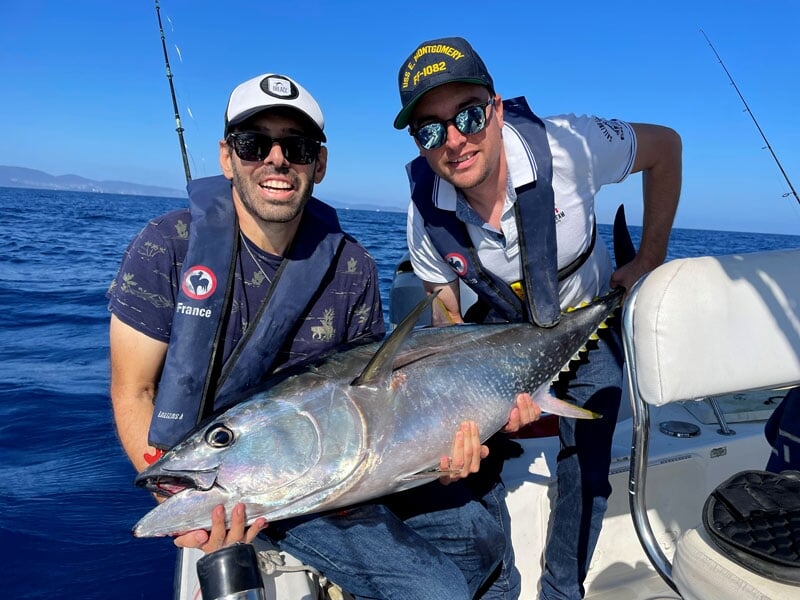 Deux hommes en tenue sportive et lunettes de soleil, tenant un poisson argenté à la forme élancée, sur un bateau en mer par temps ensoleillé
