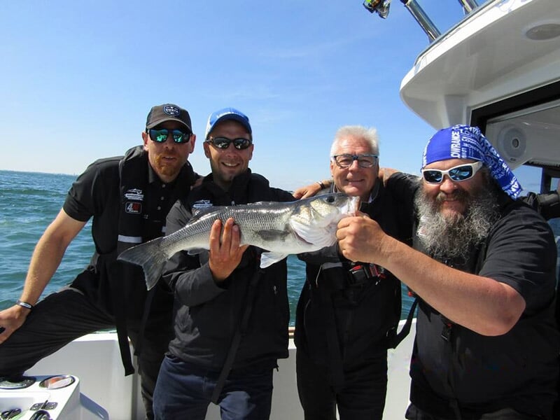 Quatre hommes souriants sur un bateau tenant un gros poisson fraîchement pêché en mer par une journée ensoleillée.