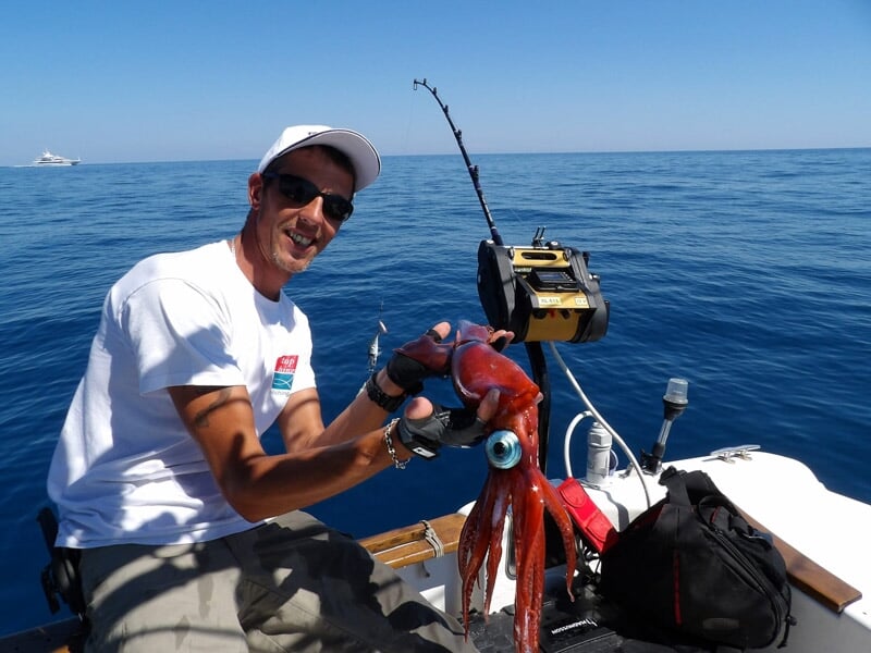 Homme souriant sur un bateau montrant un calmar rouge fraîchement pêché sous un ciel dégagé.