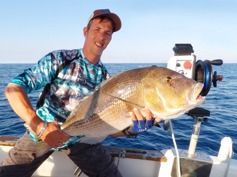 Pêcheur assis sur un bateau tenant un grand poisson beige avec des reflets dorés en mer calme.