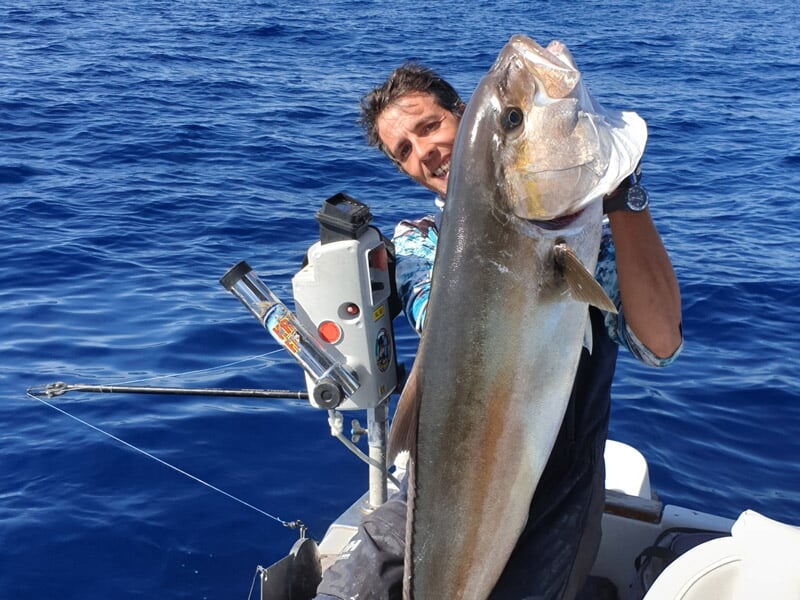 Homme sur un bateau tenant un très gros poisson argenté avec un équipement de pêche visible.