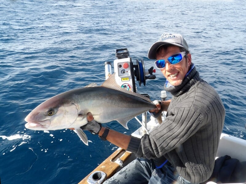 Pêcheur souriant avec des lunettes bleues tenant un poisson argenté sur un bateau en mer calme.