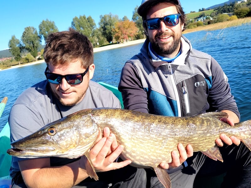 Deux pêcheurs souriants tenant un grand brochet dans un bateau sur un lac, avec un paysage paisible et un ciel bleu en arrière-plan
