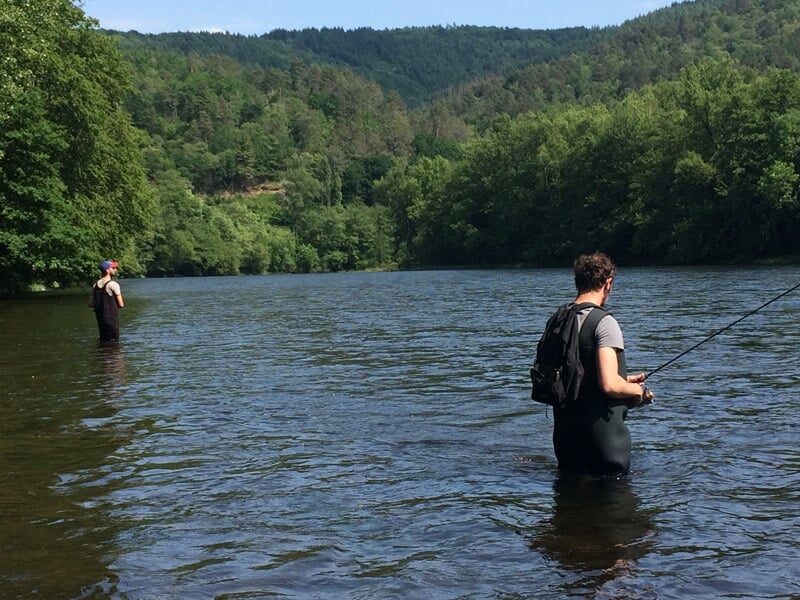 Deux pêcheurs en waders debout dans une rivière entourée de collines boisées, concentrés sur leur ligne de pêche.