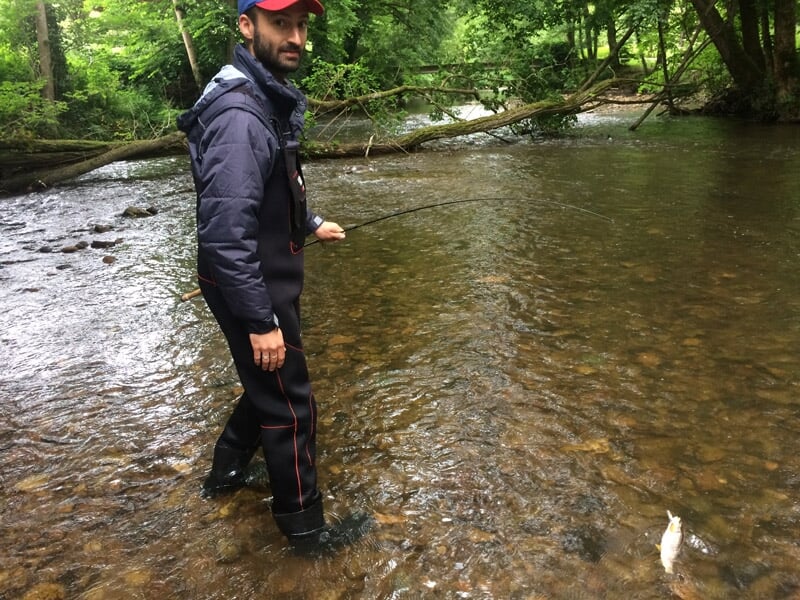 Homme en tenue de pêche debout dans une rivière peu profonde, tenant une canne à pêche avec un poisson accroché.