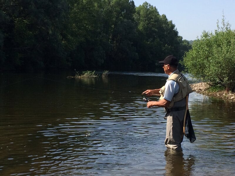 Pêcheur en waders debout dans une rivière calme, concentré sur sa ligne de pêche par une journée ensoleillée.