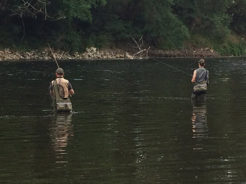 Deux pêcheurs en waders dans une rivière, pêchant côte à côte dans un environnement naturel verdoyant.