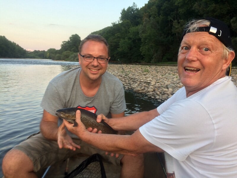 Deux hommes souriants tenant un poisson fraîchement pêché au bord d'une rivière au coucher du soleil.