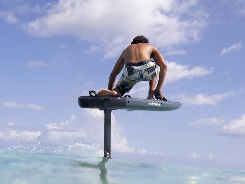Homme en maillot de bain sur un surf électrique en équilibre au-dessus de l'eau turquoise sous un ciel bleu clair.