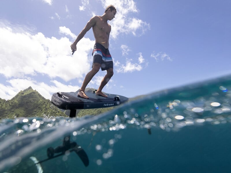 Surfeur debout sur un surf électrique glissant sur l'eau avec une montagne verte en arrière-plan sous un ciel ensoleillé.