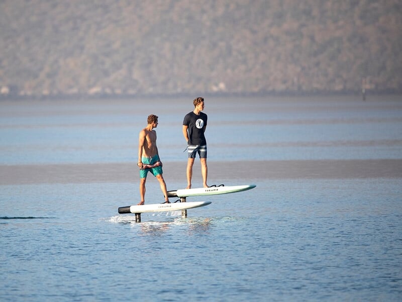 Deux hommes debout sur des surfs électriques sur une mer calme avec une côte montagneuse floue en arrière-plan.
