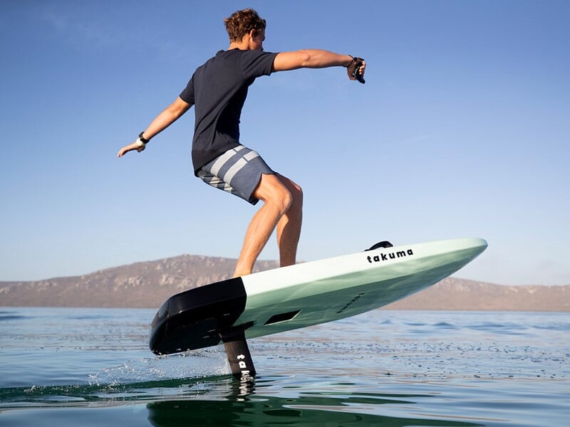 Jeune homme en short sur un surf électrique vert clair flottant au-dessus de l'eau calme avec un paysage montagneux au loin.