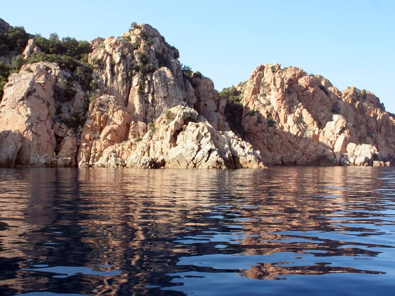 Rochers clairs se reflétant dans une mer calme avec un ciel bleu et quelques nuages légers.