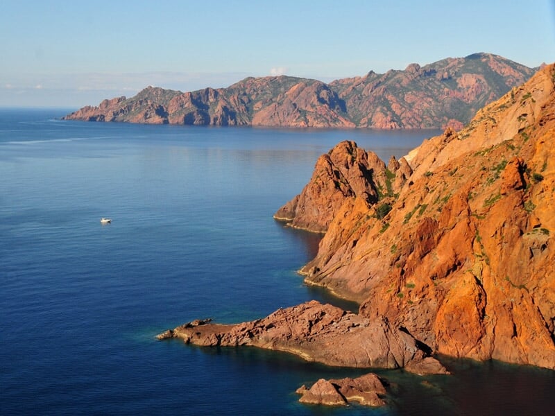 Panorama de falaises rouges s'étendant le long d'une mer bleue calme sous un ciel clair et lumineux.