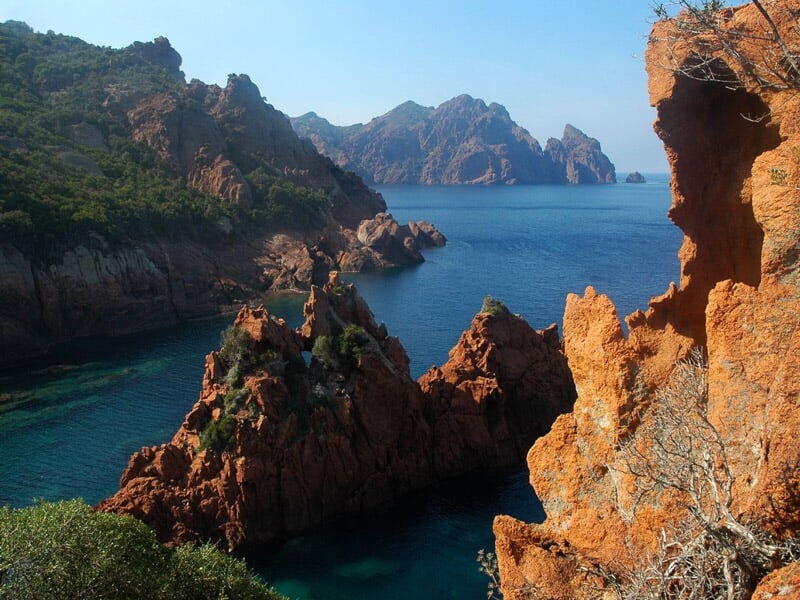 Paysage côtier avec des rochers rouges escarpés et une mer bleue profonde sous un ciel dégagé.