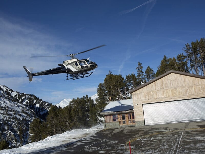 Hélicoptère volant au-dessus d'une zone enneigée avec un bâtiment en bois et des montagnes en arrière-plan.