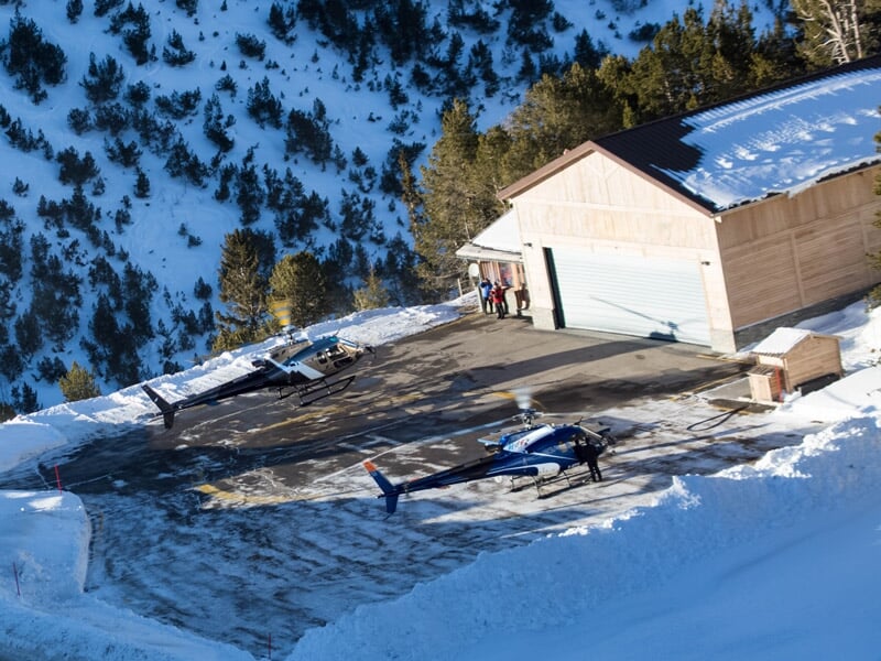 Deux hélicoptères au sol sur une plateforme enneigée près d'un bâtiment en bois entouré de montagnes.