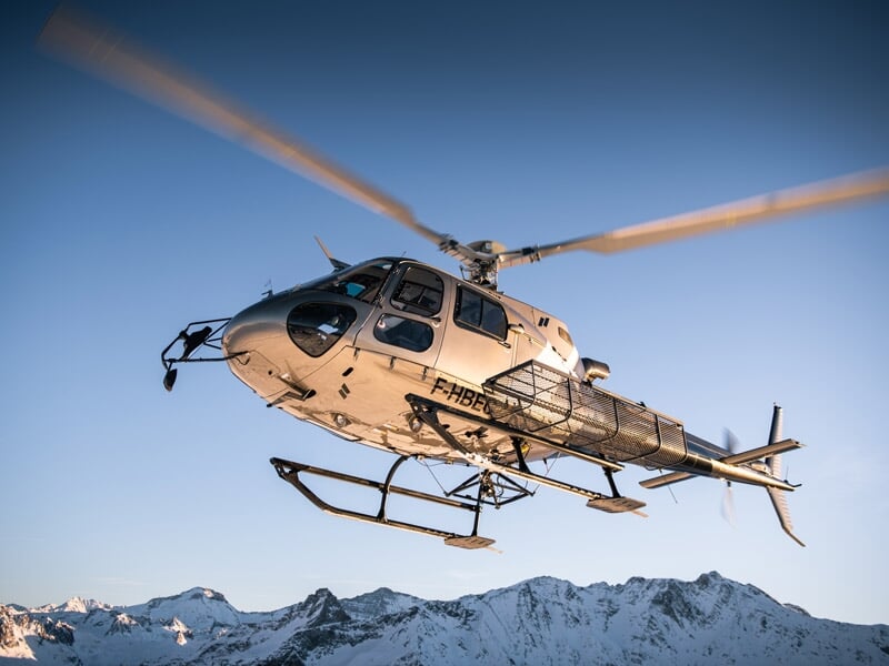 Hélicoptère en vol avec des montagnes enneigées en arrière-plan sous un ciel bleu clair.