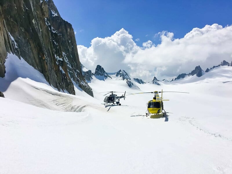 Deux hélicoptères posés sur un vaste champ de neige entouré de montagnes et de pics rocheux.
