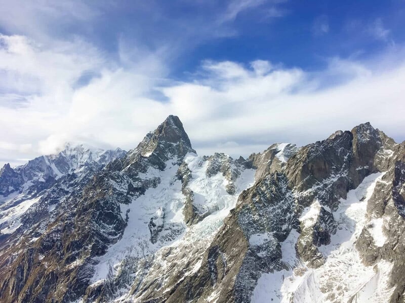 Vue aérienne de montagnes rocheuses et enneigées sous un ciel partiellement nuageux.