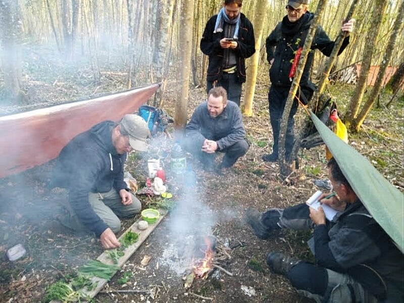 Groupe de personnes autour d'un feu de camp en forêt, préparant un repas et discutant sous un abri de fortune.