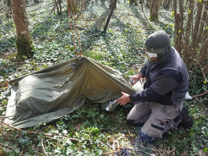 Homme installant un abri de survie en toile dans une forêt dense, entouré de végétation et de feuilles.