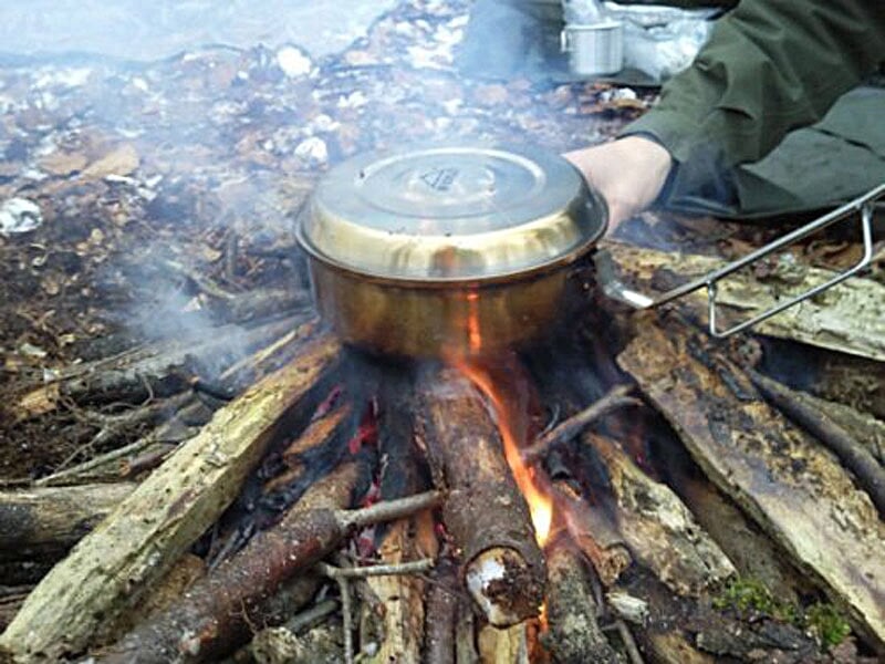 Casserole en métal posée sur un feu de camp avec des bûches enflammées dans un environnement forestier.