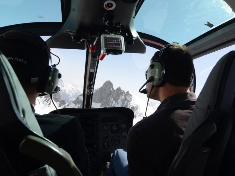 Vue intérieure d'un hélicoptère avec deux personnes portant des casques, survolant des montagnes enneigées.