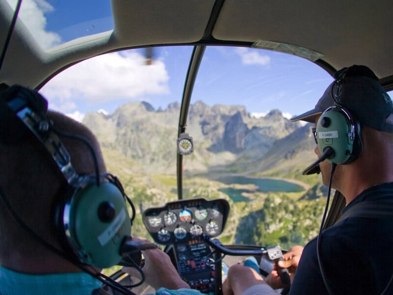 Pilote et passager dans un hélicoptère survolant un lac entouré de montagnes rocheuses sous un ciel clair.