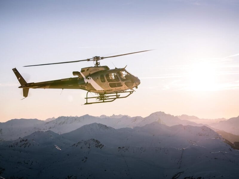 Hélicoptère beige volant au coucher du soleil au-dessus de montagnes enneigées avec un ciel aux teintes chaudes.