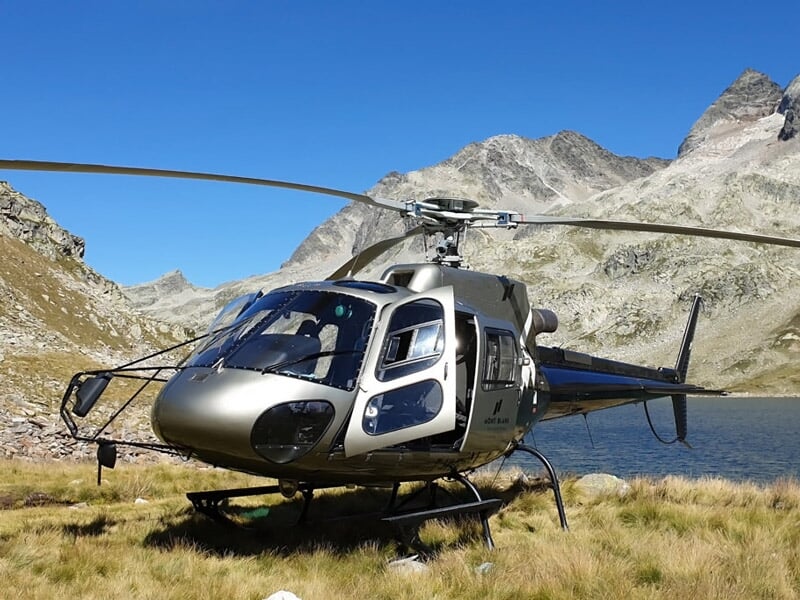 Hélicoptère gris posé près d'un lac de montagne avec des sommets rocheux en arrière-plan sous un ciel bleu.