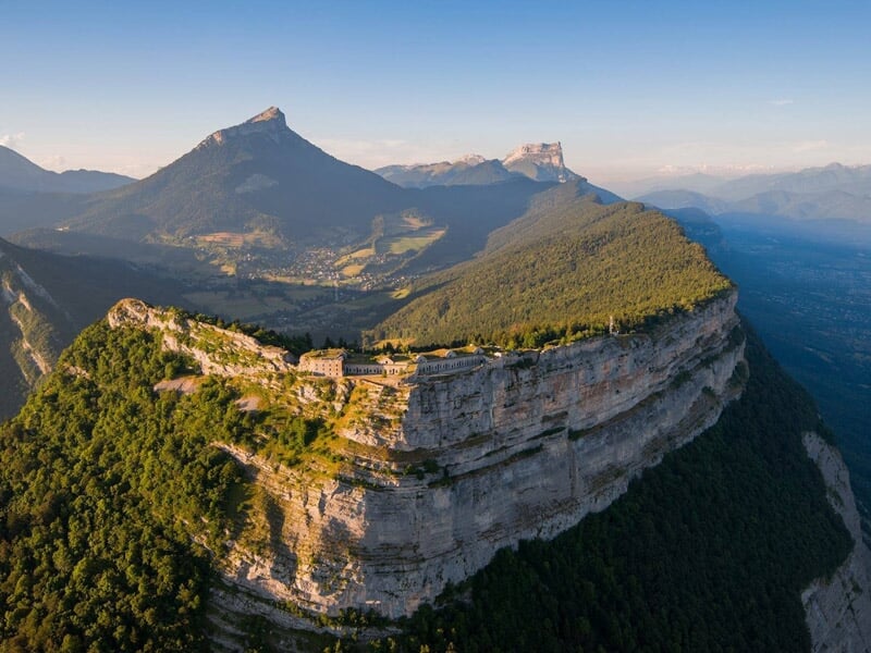 Vue aérienne d'une falaise escarpée avec une forteresse au sommet, entourée de montagnes et de forêts vertes.